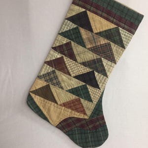 May include: A green, brown, and beige patchwork Christmas stocking with a quilted design. The stocking is made of various plaid fabrics and features a geometric pattern of triangles.