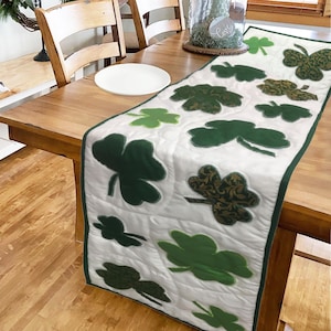 May include: White quilted table runner with green shamrock designs. The shamrocks are in various sizes and shades of green. The runner is perfect for a St. Patrick's Day celebration.