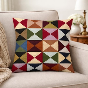 May include: Square throw pillow with a geometric patchwork design. The pillow features a variety of colors including red, beige, black, green, and brown. The pillow is placed on a beige armchair.