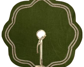 Puede incluir: Una falda verde para árbol de Navidad con borde festoneado y ribete plateado. La falda tiene una abertura central para el tronco del árbol y está sostenida por un soporte plateado.