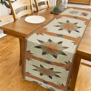 May include: A rectangular table runner with a star pattern in shades of cream, brown, and rust. The runner features large, eight-pointed stars and geometric accents. The design is reminiscent of a vintage quilt.