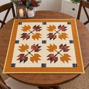 May include: A square fabric table topper featuring a repeating pattern of four maple leaf motifs. The leaves are rendered in shades of orange, brown, and plaid, set against a cream background with small dark blue plaid squares and an orange border.