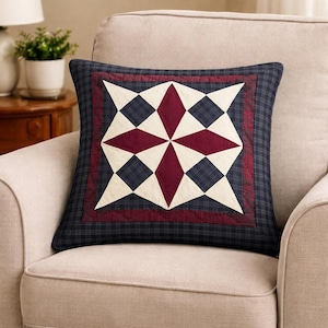 Könnte beinhalten: Quadratisches Zierkissen mit Sternmuster. Das Kissen hat einen marineblauen Karomuster-Rand und ein zentrales Muster aus cremefarbenen, weinroten und marineblauen geometrischen Formen. Das Kissen liegt auf einem beigen Sessel.