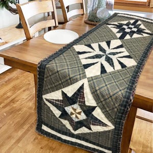 May include: A quilted table runner with a plaid pattern in shades of green, blue, and brown. The runner features three large star-shaped designs in white, navy, and brown. The runner is displayed on a wooden table.