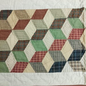May include: A quilted table runner featuring a geometric pattern of 3D cubes. The cubes are composed of various fabrics in shades of red, green, beige, and blue, with a white border and brown trim.