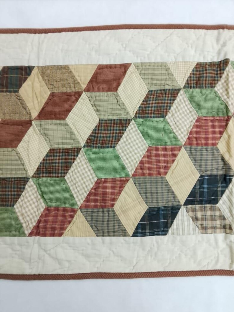 May include: A quilted table runner featuring a geometric pattern of 3D cubes. The cubes are composed of various fabrics in shades of red, green, beige, and blue, with a white border and brown trim.