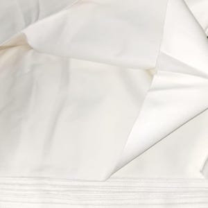 May include: A close-up shot of folded white fabric, possibly cotton or linen. The fabric is smooth and appears to be new, with visible folds and creases. The image highlights the texture and pristine condition of the material, suitable for sewing or crafting projects.