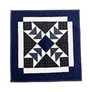 May include: A square quilt with a navy blue border and a central starburst design. The starburst features triangles in shades of gray and white, set against a black and gray plaid pattern. The quilt is machine-stitched.