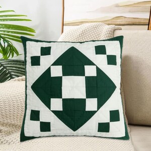 Puede incluir: Cojín cuadrado con un diseño geométrico. El cojín presenta una forma de diamante central en verde oscuro, rodeada de cuadrados blancos y un borde verde. El cojín está sobre un sofá beige.
