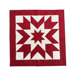 May include: A square, quilted textile featuring a central starburst design in white and red. The star is surrounded by white and red triangles, with a solid red border. The quilt is likely handmade.