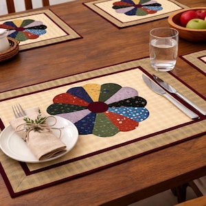 May include: A wooden table set with four placemats, each with a colourful, flower-like design. A plate with a napkin and silverware, a glass of water, and a bowl of fruit are also on the table. The placemats have a beige background with a burgundy border.
