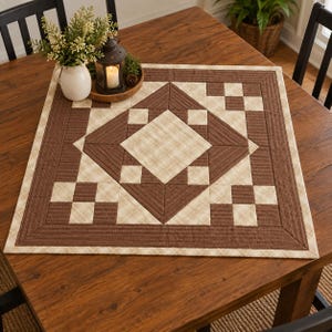 Op de afbeelding: Een vierkant stoffen tafelkleed met een geometrisch patchworkontwerp in bruin- en crèmetinten. Het patroon bestaat uit een centrale ruit omringd door kleinere vierkanten en driehoeken, afgewerkt met een brede, doorgestikte bruine rand.