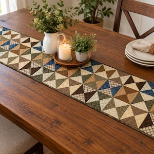 Puede incluir: Un camino de mesa acolchado rectangular con un patrón geométrico de triángulos en tonos marrón, bronceado, crema, verde oliva y azul marino. La tela muestra una mezcla de colores sólidos y pequeños estampados de cuadros con costuras visibles.