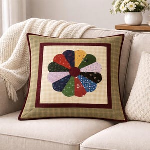 May include: Square decorative pillow with a colorful flower design. The flower has petals in various colors, including red, yellow, green, and blue. The pillow has a cream-colored background with a plaid pattern and a dark red border. The pillow is on a beige sofa.