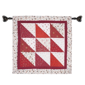 May include: A red and white patchwork quilt wall hanging with a floral print border. The quilt is made of various fabrics with different patterns and is hung with black metal hooks.