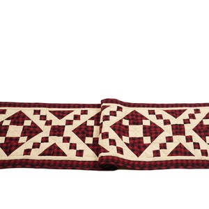 May include: A red and white checkered table runner with a geometric pattern. The runner is made of quilted fabric and has a fringed edge.