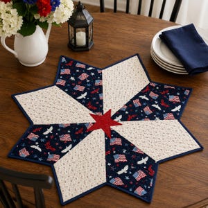 May include: A star-shaped quilted table topper with a patriotic theme. The topper features alternating sections of navy blue fabric with American flags and white fabric with small red floral patterns. A red star is in the center.