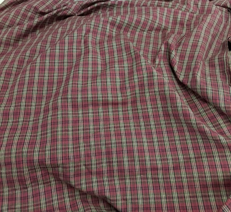 May include: Close-up of a fabric with a plaid pattern. The fabric features a burgundy base with intersecting lines in black and light green, creating a classic plaid design. The fabric is slightly wrinkled.