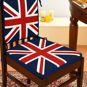 Op de afbeelding: Een houten stoel met kussens met het ontwerp van de Britse vlag. De kussens hebben een marineblauwe achtergrond met het rood-witte kruis. De stoel staat op een patroonkleed, met een tafel en fruit op de achtergrond.