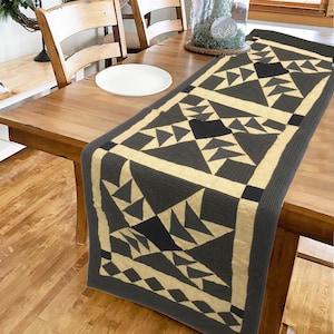 Op de afbeelding: Een zwarte en crèmekleurige gestikte tafelrunner met een geometrisch patroon. De runner heeft in elke sectie een ruitvorm in het midden, omringd door driehoeken.