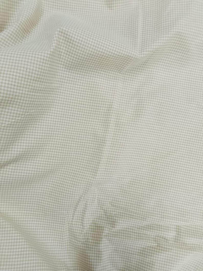 May include: Close-up of a light beige fabric with a small, evenly spaced checkered pattern. The fabric is slightly wrinkled, creating shadows and variations in tone. The overall appearance suggests a soft, woven material, possibly cotton or linen.