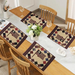 May include: A wooden table set with patriotic-themed placemats and a runner. The placemats and runner feature a quilted design with the American flag and diamond patterns in red, white, blue, and tan. The table is set with wine glasses and a vase of white flowers.