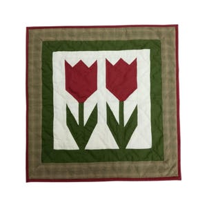 May include: A square quilted wall hanging featuring two red tulip flower designs on a white background. The tulips have green stems and leaves. The design is framed by a green border, a brown plaid border, and a red border.