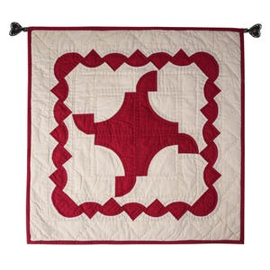 May include: A red and white quilted wall hanging with a geometric design. The design features a large red cross shape in the center, surrounded by a white border and a red scalloped edge.