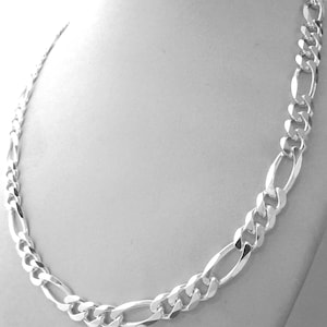 May include: A silver Figaro chain necklace displayed on a mannequin. The necklace features a pattern of three small circular links followed by one elongated oval link. The chain is made of a shiny, reflective metal.
