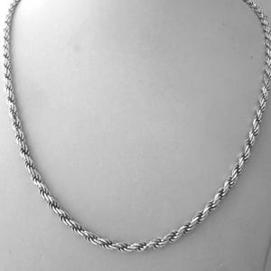 以下が含まれることがあります： シルバーカラーのロープチェーンネックレス。白いマネキンに飾られています。チェーンは、ロープのような質感を作り出す、しっかりと織られたリンクでできています。ネックレスはクラシックなジュエリーです。