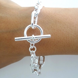 Peut inclure: Un bracelet argenté avec un fermoir à bascule. Le bracelet présente un motif de chaîne et une fermeture à bascule circulaire. Le bracelet est présenté sur un poignet.