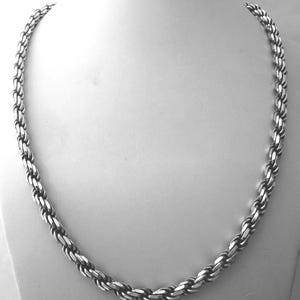 May include: A silver-colored rope chain necklace displayed on a white mannequin. The chain is made of interlocking oval links, creating a textured, braided appearance. The necklace is a classic jewelry piece.