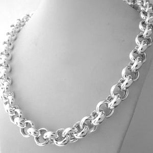 May include: A silver-colored chain necklace with large, rounded links. The necklace is displayed on a white mannequin bust, showcasing its design and texture. The links are closely connected, creating a bold and eye-catching accessory.
