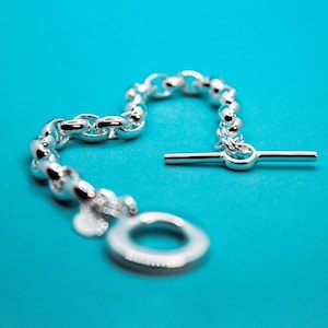 Peut inclure: Un bracelet en argent avec un fermoir à glissière et un charme rond. Le bracelet est sur un fond bleu.