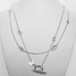 May include: A silver chain necklace with a toggle clasp. The chain is made of two strands of silver links, one thicker than the other. The necklace has a small silver charm with a design on it.