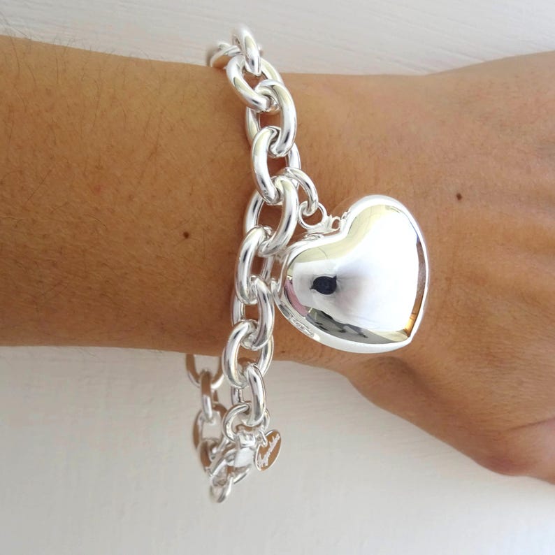 May include: A silver-colored bracelet with a large heart-shaped charm. The bracelet features a chunky chain design and a polished heart pendant. The bracelet is shown on a person's wrist.