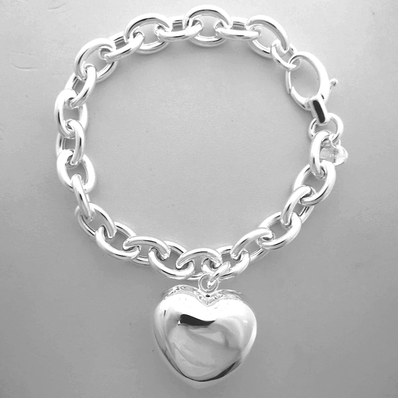 May include: A silver-colored bracelet with a large heart pendant. The bracelet features a chunky chain design with an oval clasp. The heart pendant is smooth and reflective, adding a touch of elegance to the jewelry.