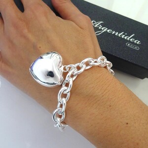 May include: A silver bracelet with a large heart-shaped charm. The bracelet features a chunky chain design. The heart charm is polished and reflective. The bracelet is displayed on a wrist, with a black box in the background.