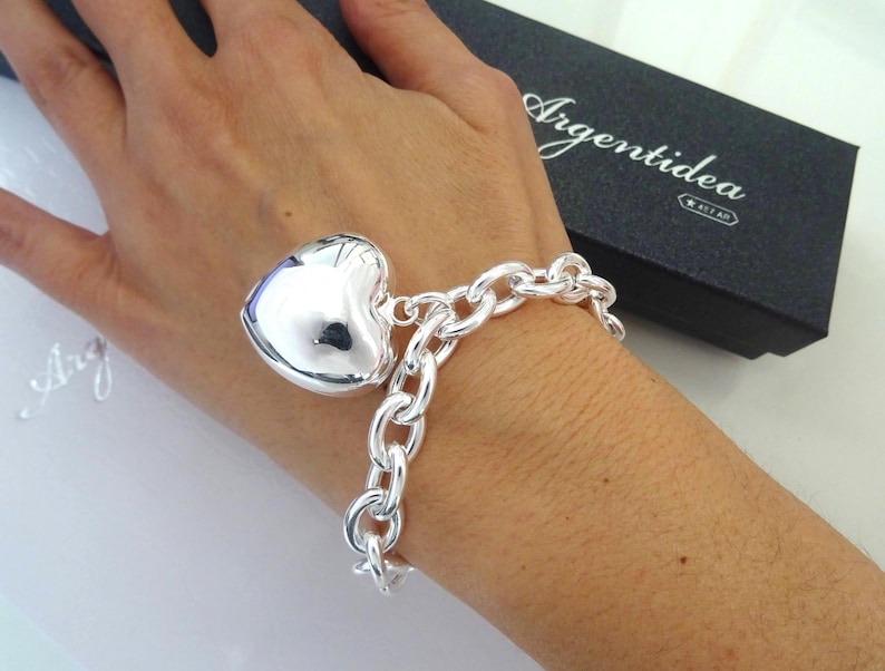 May include: A silver bracelet with a large heart-shaped charm. The bracelet features a chunky chain design. The heart charm is polished and reflective. The bracelet is displayed on a wrist, with a black box in the background.