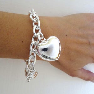 May include: A silver-colored bracelet with a large, heart-shaped charm. The bracelet features a chunky chain design, and the heart charm is smooth and reflective. Additional smaller heart charms dangle from the chain.