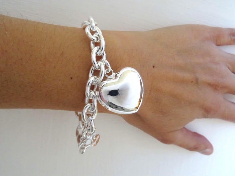 May include: A silver-colored bracelet with a large, heart-shaped charm. The bracelet features a chunky chain design, and the heart charm is smooth and reflective. Additional smaller heart charms dangle from the chain.