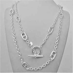 May include: A silver-toned necklace with a double-strand design. The necklace features a toggle clasp and a chain with oval-shaped links. The jewelry is displayed on a mannequin.