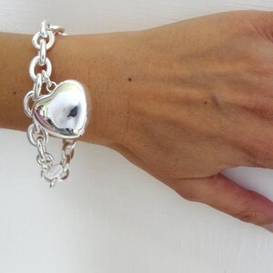 May include: A silver bracelet with a large heart-shaped charm. The bracelet features a chain link design and is worn on a wrist. The heart charm is polished and reflective, catching the light. The bracelet is a piece of jewelry.