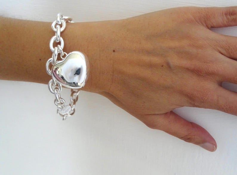 May include: A silver bracelet with a large heart-shaped charm. The bracelet features a chain link design and is worn on a wrist. The heart charm is polished and reflective, catching the light. The bracelet is a piece of jewelry.