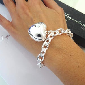 May include: A silver bracelet with a large heart charm. The bracelet features a chunky chain design and a polished heart pendant. The bracelet is displayed on a person's wrist, with a black box in the background that reads "Argentidea".