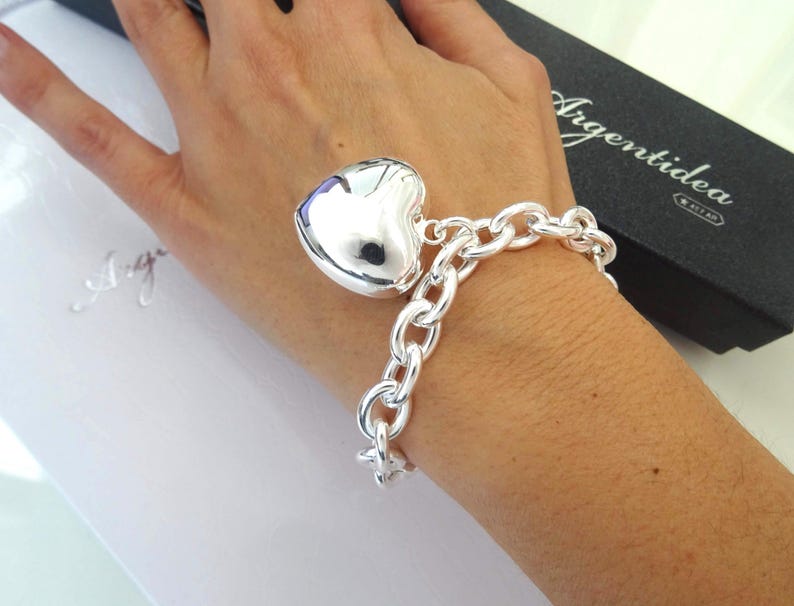 May include: A silver bracelet with a large heart charm. The bracelet features a chunky chain design and a polished heart pendant. The bracelet is displayed on a person's wrist, with a black box in the background that reads "Argentidea".