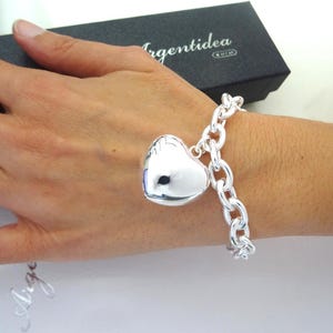 May include: A silver bracelet with a large heart-shaped charm. The bracelet features a chunky chain design. The heart charm is smooth and reflective. The bracelet is displayed on a wrist.