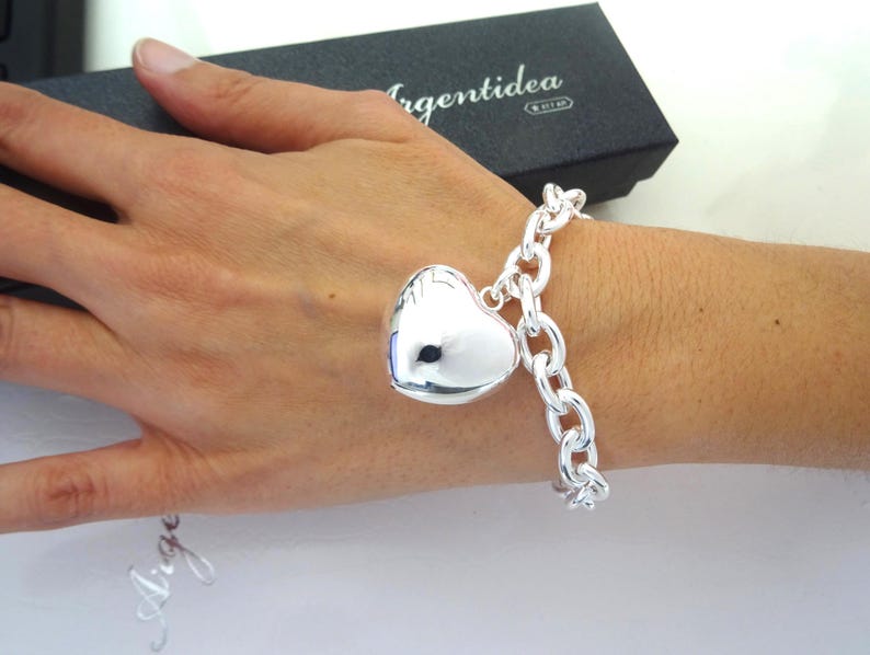 May include: A silver bracelet with a large heart-shaped charm. The bracelet features a chunky chain design. The heart charm is smooth and reflective. The bracelet is displayed on a wrist.