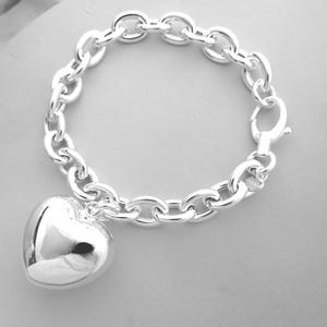 May include: A silver bracelet with a large heart charm. The bracelet features a chain link design and a lobster clasp closure. The heart charm is smooth and reflective, adding a touch of elegance to the jewelry.