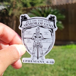 May include: A white sticker with a black outline of a knight in armor. The knight is holding a sword and is surrounded by a shield with the words "Put on the full armor of God" and "Ephesians 6:10".